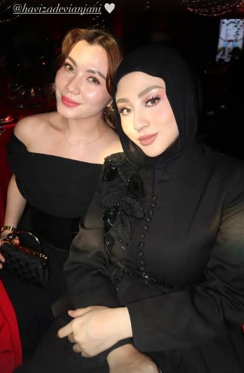 Dua wanita berbusana elegan berpose bersama di acara resepsi pernikahan dengan latar pencahayaan merah yang hangat.