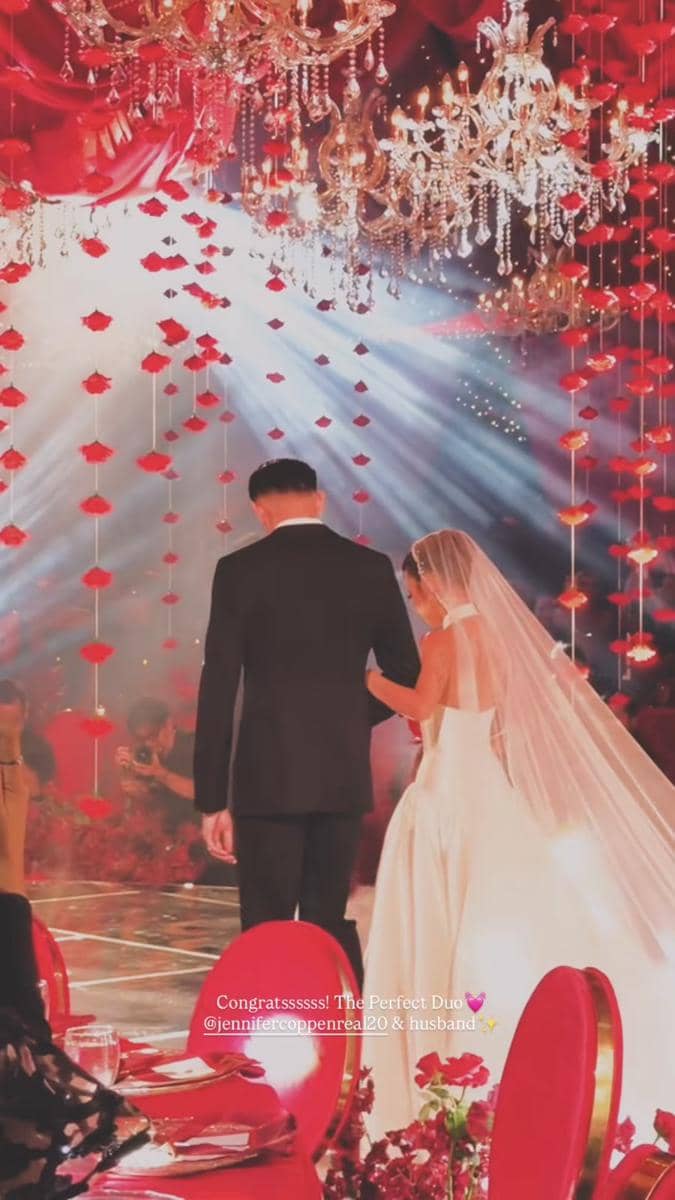 Pasangan pengantin berjalan di bawah lampu gantung kristal dan dekorasi bunga merah dalam resepsi pernikahan yang mewah.