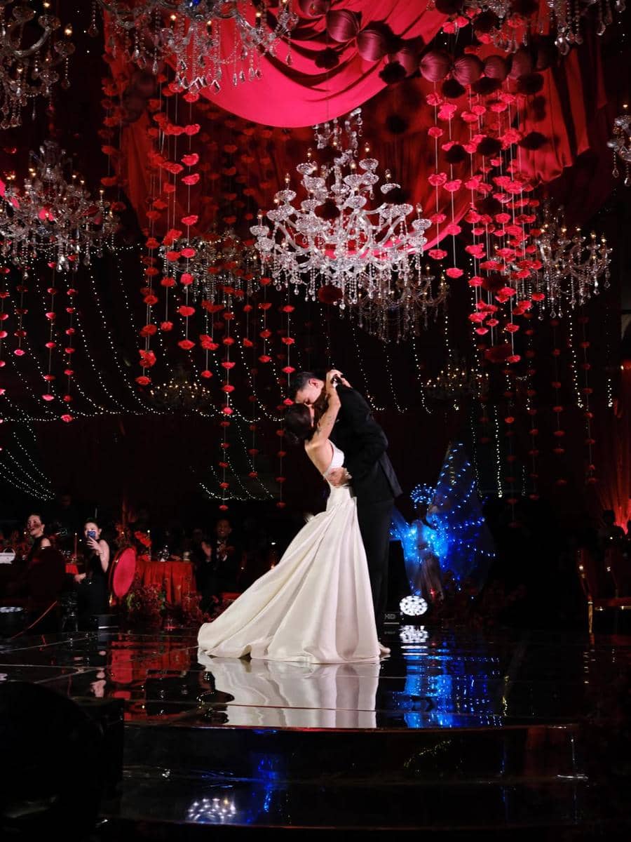 Pasangan pengantin menari di lantai dansa dengan dekorasi mewah bernuansa merah dan lampu gantung kristal di resepsi pernikahan.