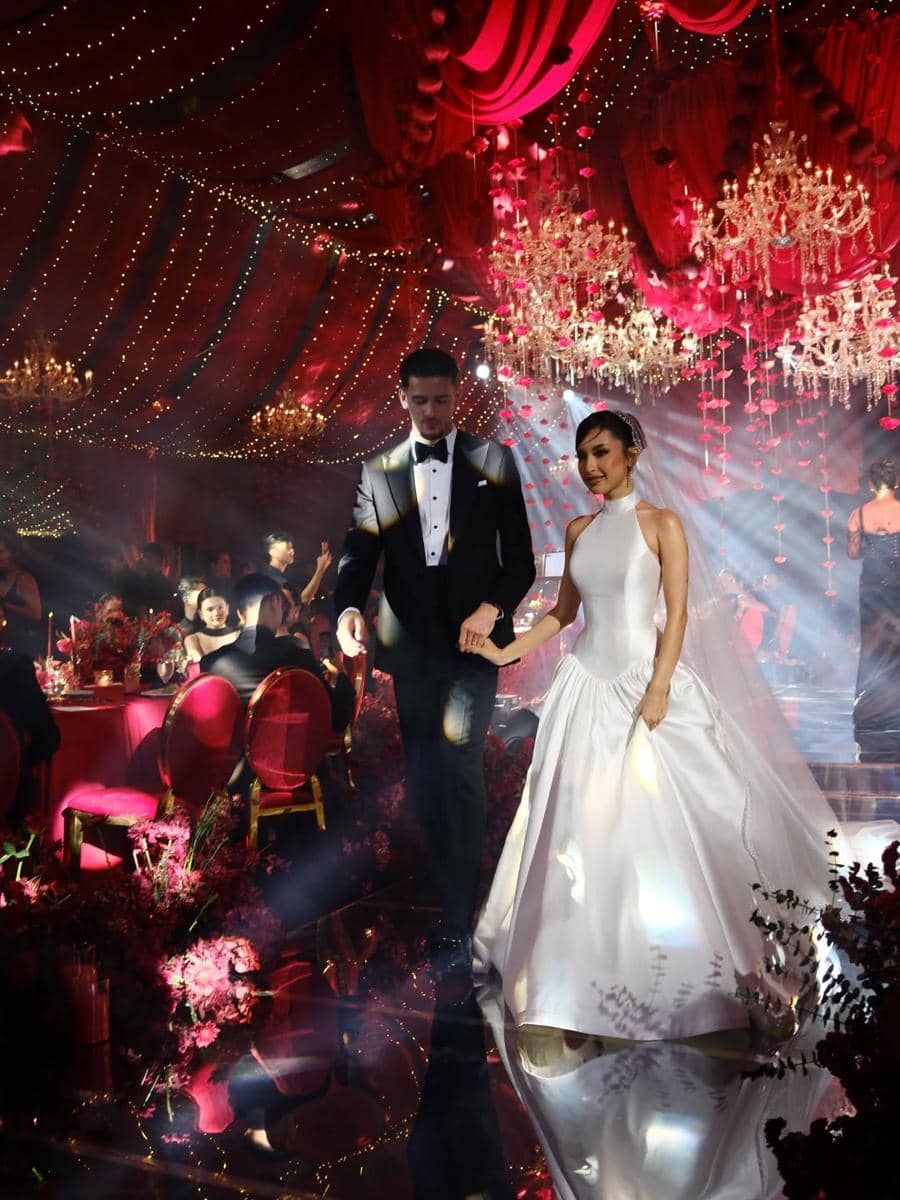 Pasangan pengantin berjalan di tengah dekorasi mewah dengan lampu gantung kristal dan nuansa merah pada resepsi pernikahan.