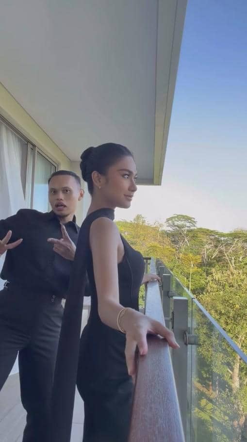Dua tamu berpakaian hitam berpose di balkon dengan latar pepohonan hijau saat menghadiri resepsi pernikahan.