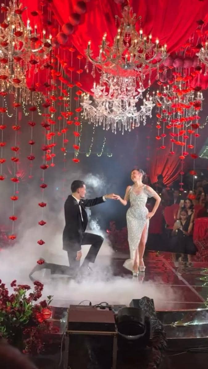 Pasangan mengenakan busana formal menari di panggung berhias lampu gantung kristal dan dekorasi bunga merah dengan nuansa romantis.