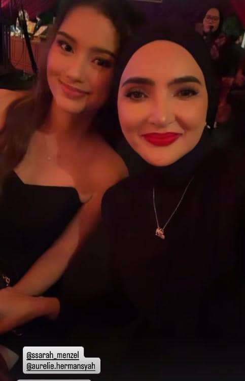 Dua wanita berpose bersama mengenakan busana elegan berwarna hitam di acara resepsi pernikahan dengan pencahayaan hangat.