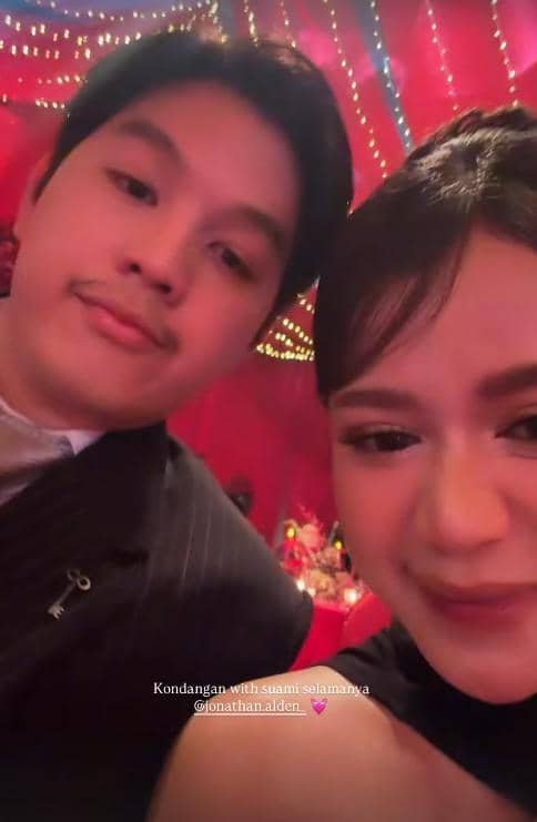 Sepasang tamu berpose bersama di resepsi pernikahan Jennifer Coppen dan Justin dengan latar dekorasi merah dan lampu hias.