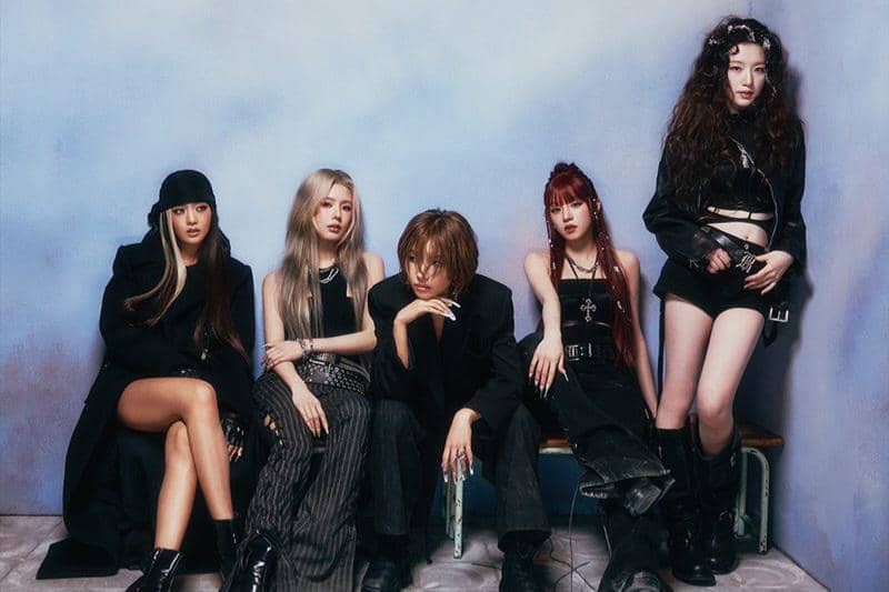 Lima anggota grup musik perempuan berpose dengan busana bergaya edgy dan modern di depan latar berwarna biru lembut.