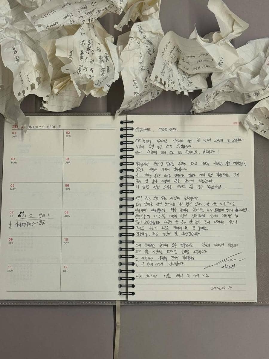 Surat tulisan tangan dalam bahasa Korea di buku catatan spiral dengan beberapa kertas kusut di sekelilingnya di atas meja.