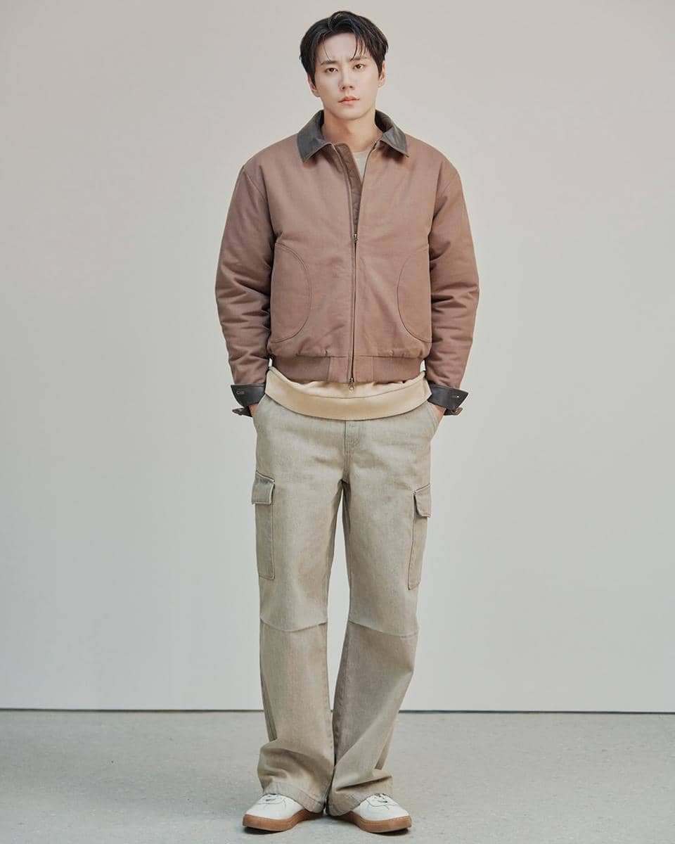 Seorang pria berdiri mengenakan jaket cokelat muda, celana kargo krem, dan sepatu putih di ruangan dengan latar polos.