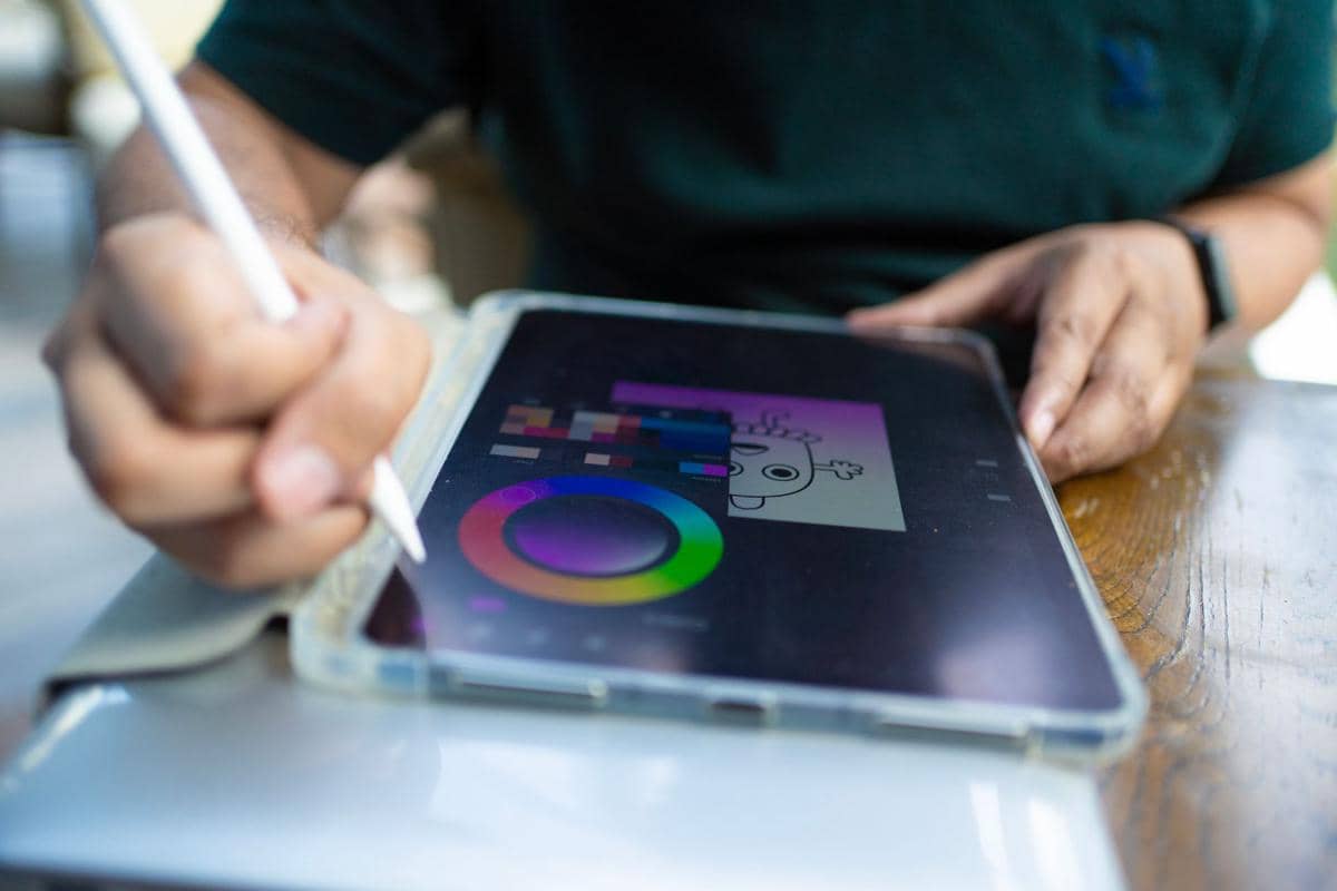 Seseorang menggunakan tablet dengan stylus di atas meja, menggambarkan aktivitas digital dan pekerjaan kreatif.