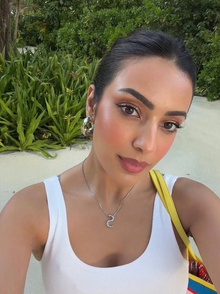 Seorang wanita berpose selfie di luar ruangan dengan latar belakang tanaman hijau, mengenakan tank top putih dan kalung perak.