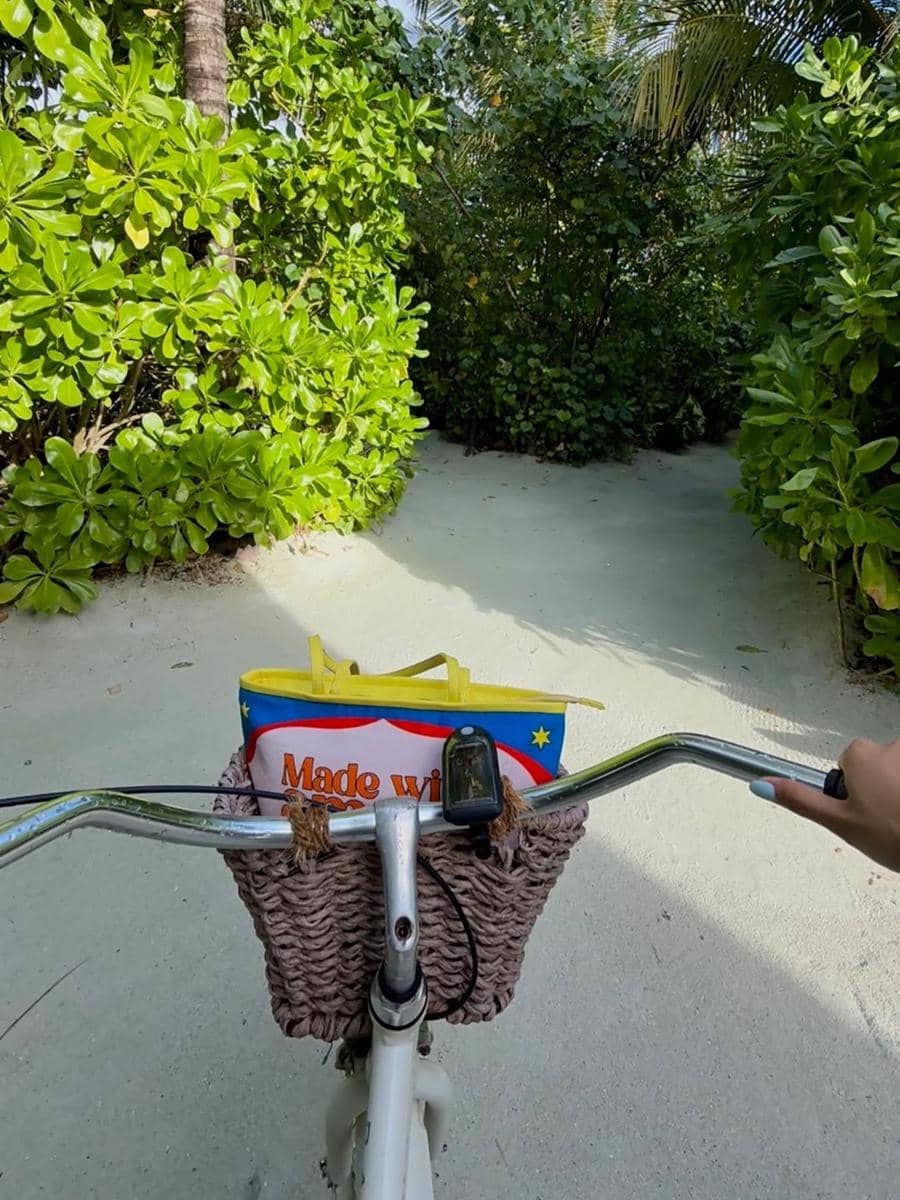 Seseorang mengendarai sepeda putih dengan keranjang anyaman di jalan berpasir yang dikelilingi pepohonan hijau tropis.