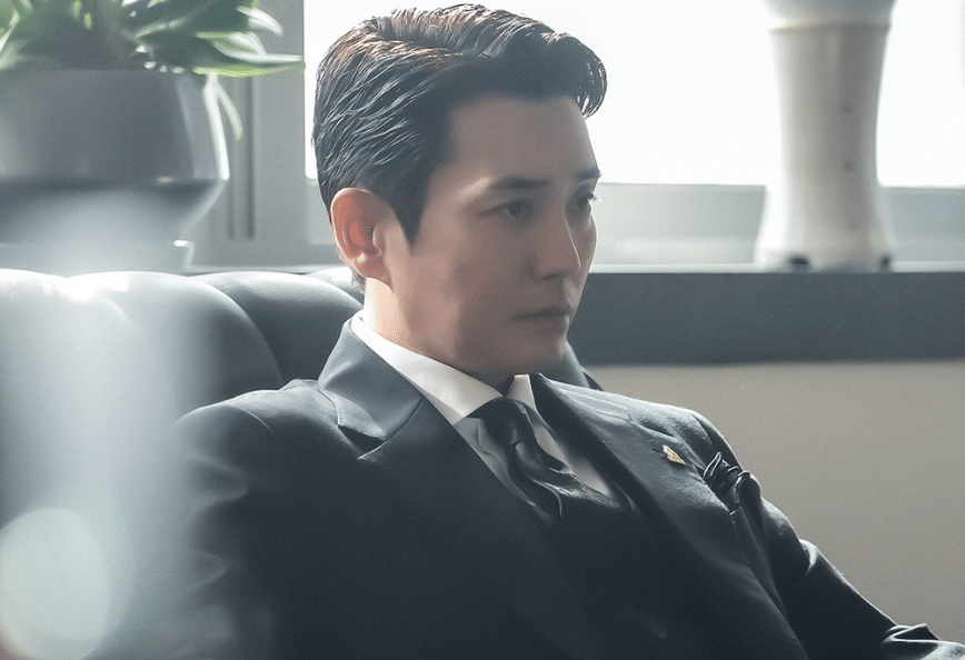Seorang pria mengenakan setelan jas hitam duduk di sofa dengan ekspresi serius dalam cuplikan drama Korea Agent Kim Reactivated.