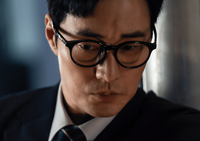 Seorang pria berkacamata mengenakan setelan jas hitam dan dasi tampak serius dalam cuplikan drama Korea Agent Kim Reactivated.