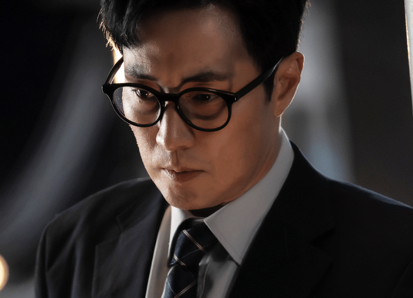 Seorang pria berkacamata mengenakan setelan jas dan dasi tampak serius dalam cuplikan drama Korea Agent Kim Reactivated.