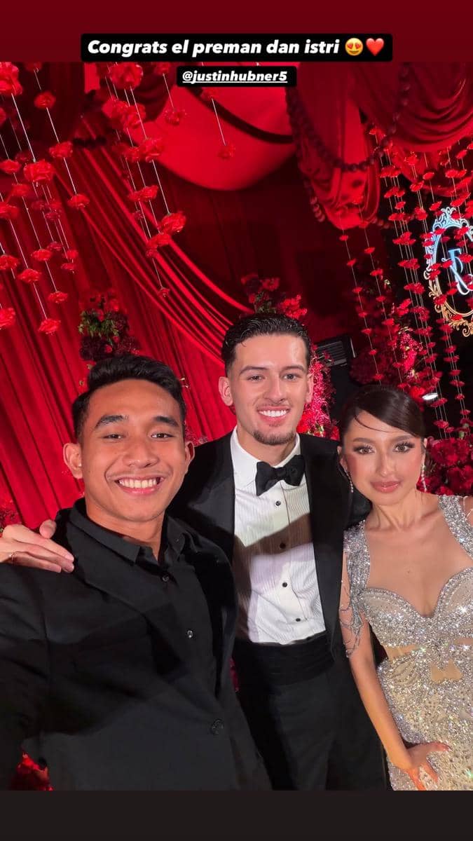 Seorang pemain timnas berfoto bersama pasangan pengantin di resepsi pernikahan dengan dekorasi merah dan bunga mawar.