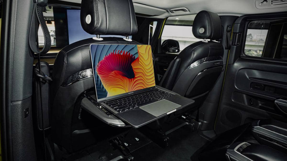 Laptop terbuka diletakkan di meja lipat belakang kursi mobil dengan interior kulit hitam yang elegan dan modern.