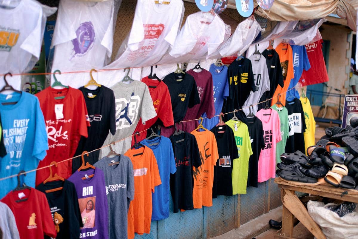 Deretan kaus berwarna-warni dengan berbagai desain dan tulisan digantung di tali jemuran di kios penjual pakaian pinggir jalan.