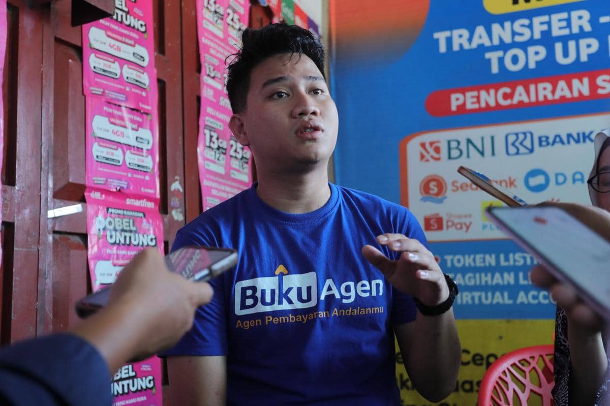 Seorang pria mengenakan kaus biru bertuliskan Buku Agen sedang berbicara kepada beberapa orang yang memegang ponsel di Deli Serdang.
