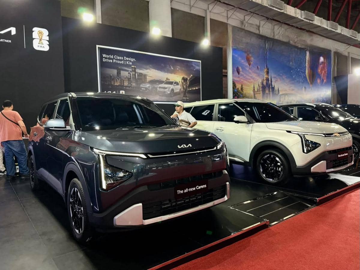Dua mobil Kia Carens dan Sonet dipamerkan di sebuah ajang otomotif dalam ruangan dengan latar belakang booth bertema FIFA.