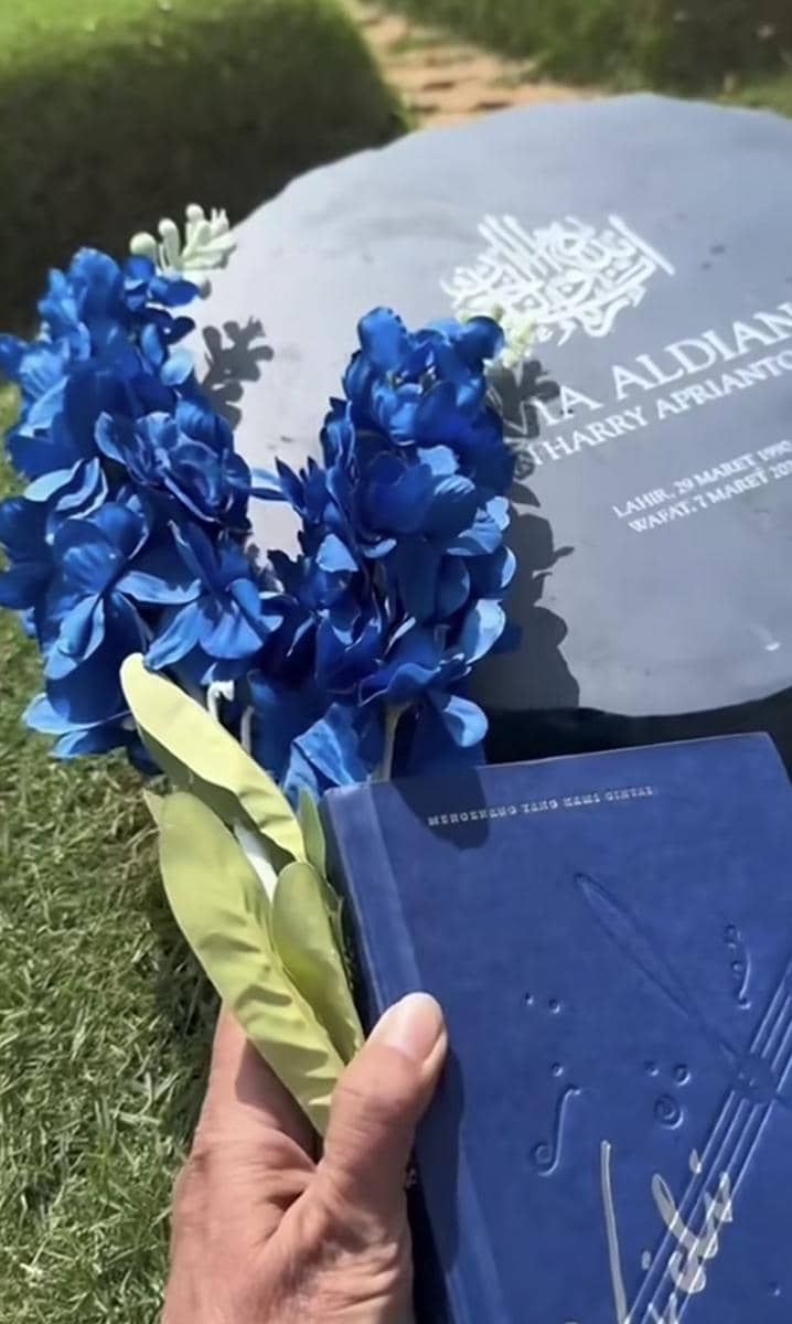 Tangan memegang bunga biru dan buku berwarna biru di atas nisan bertuliskan nama almarhum di area pemakaman yang hijau.