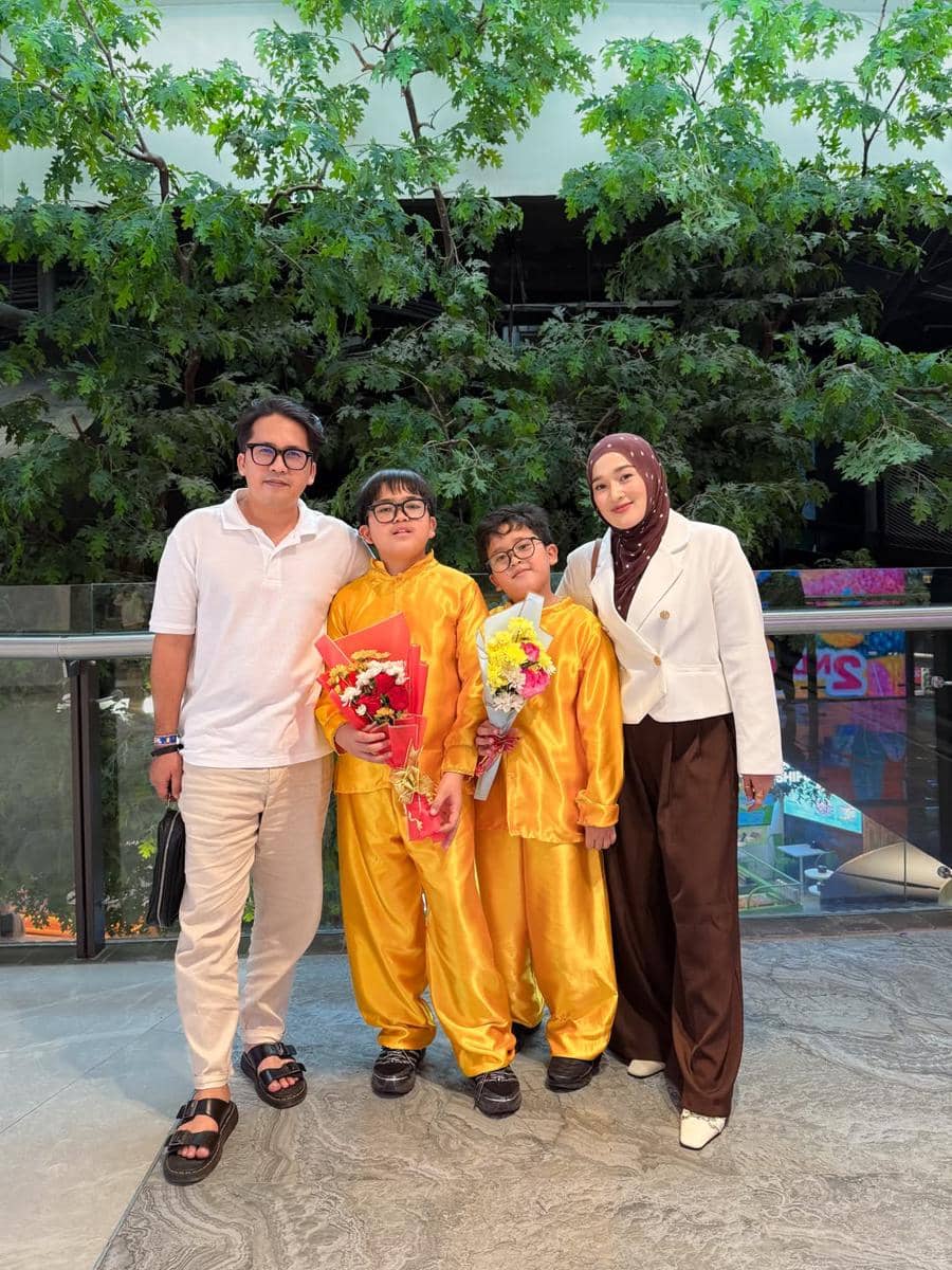 Dua orang dewasa berpose bersama dua anak mengenakan pakaian wisuda berwarna kuning sambil memegang buket bunga di area terbuka dengan latar pepohonan.