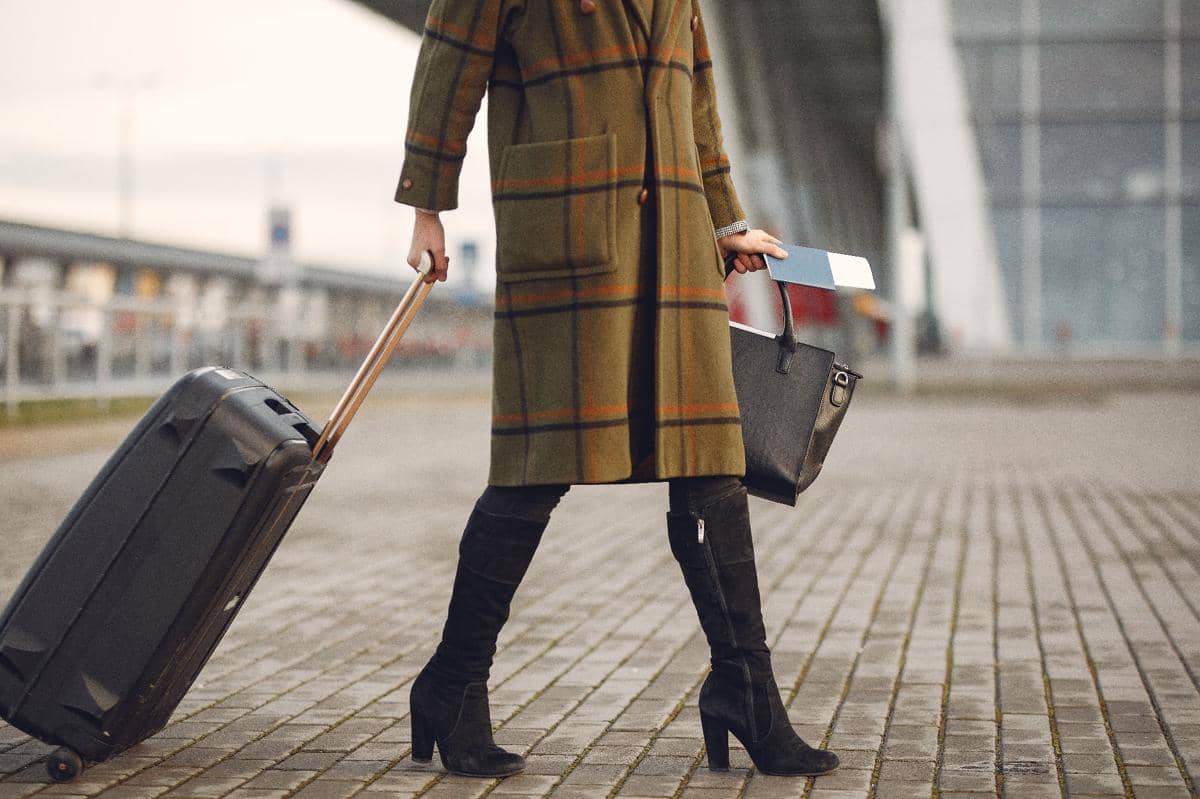 Seorang wanita mengenakan mantel panjang dan sepatu bot hitam menarik koper di trotoar dekat bangunan modern.