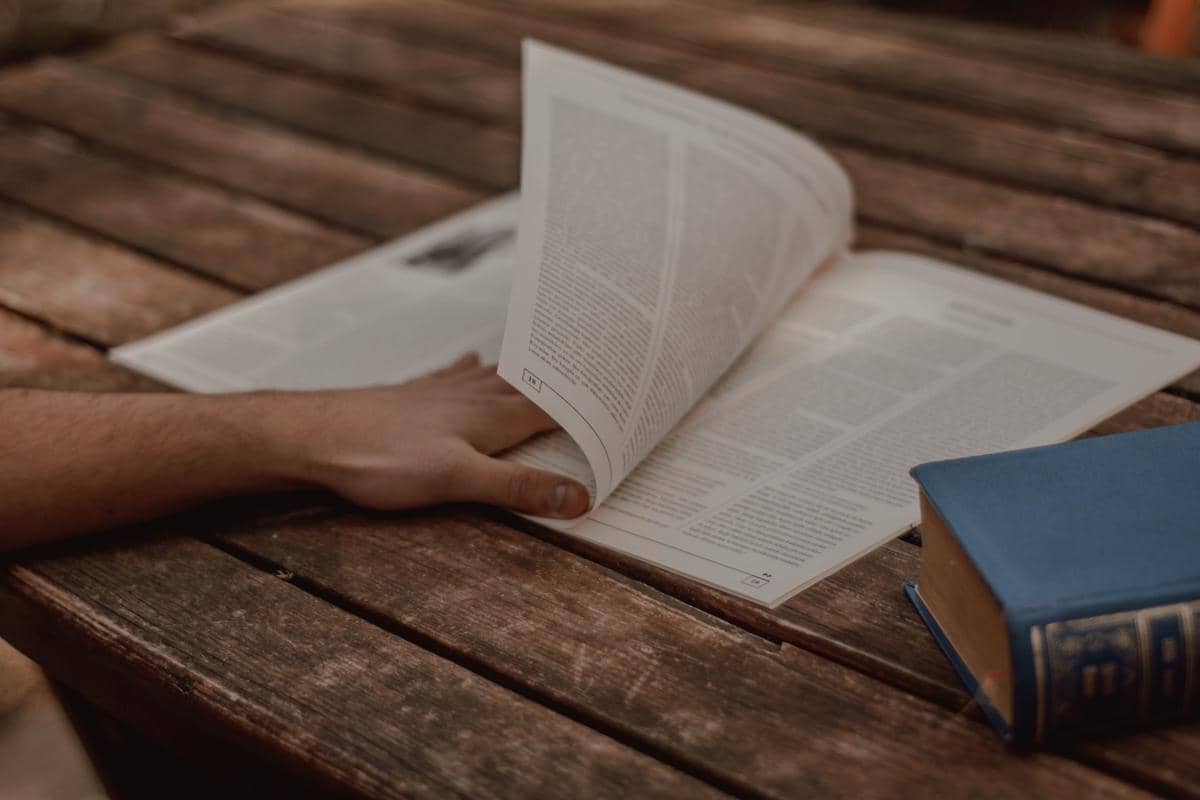 Tangan seseorang membalik halaman buku di atas meja kayu dengan sebuah buku tebal berwarna biru di sampingnya.