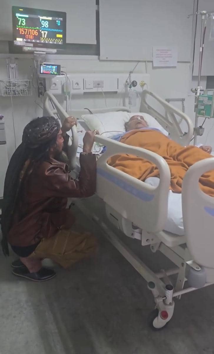 Seorang pria duduk di samping tempat tidur rumah sakit sambil memegang tangan pasien yang terbaring dengan selimut oranye.