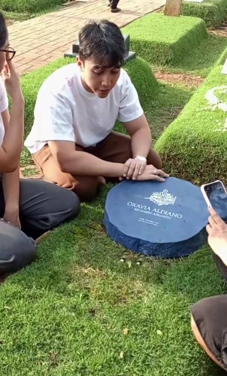 Seorang pria duduk di samping nisan berwarna biru di area pemakaman, mengenang seseorang yang telah berpulang bersama beberapa orang lain.