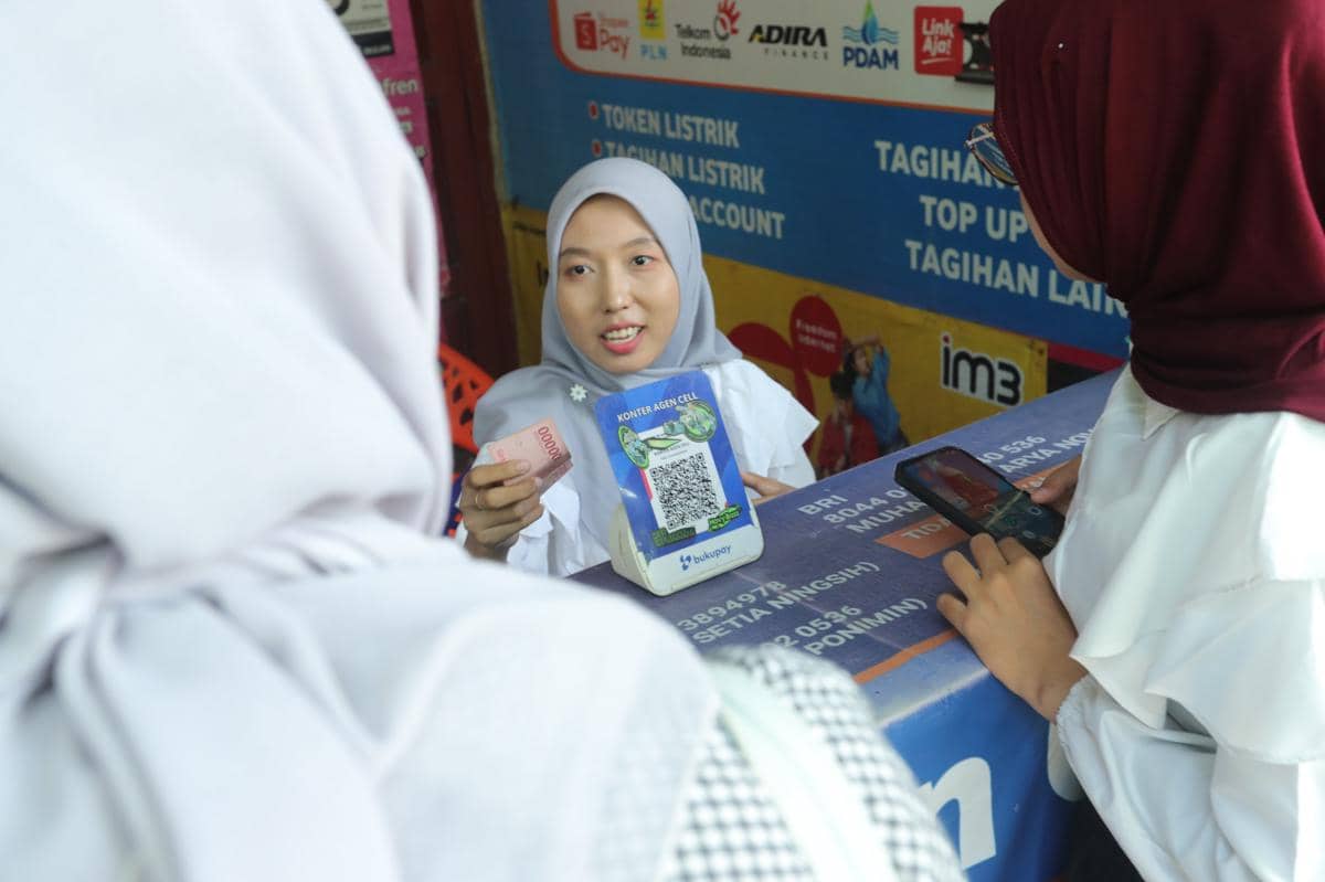 Seorang pegawai merchant lokal di Deli Serdang melayani pelanggan yang bertransaksi menggunakan uang tunai dan ponsel di meja kasir.