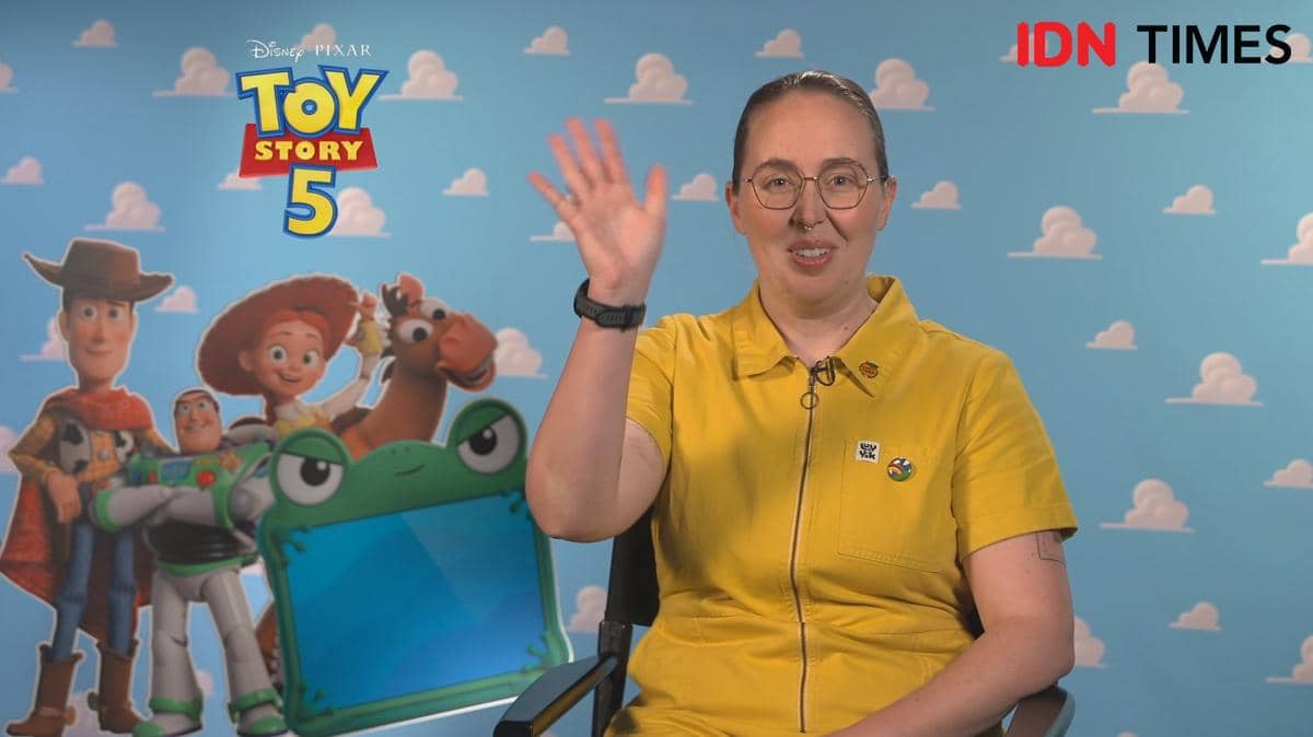 Seorang perempuan mengenakan pakaian kuning melambaikan tangan saat wawancara virtual dengan latar belakang logo Toy Story 5 dan karakter Pixar.