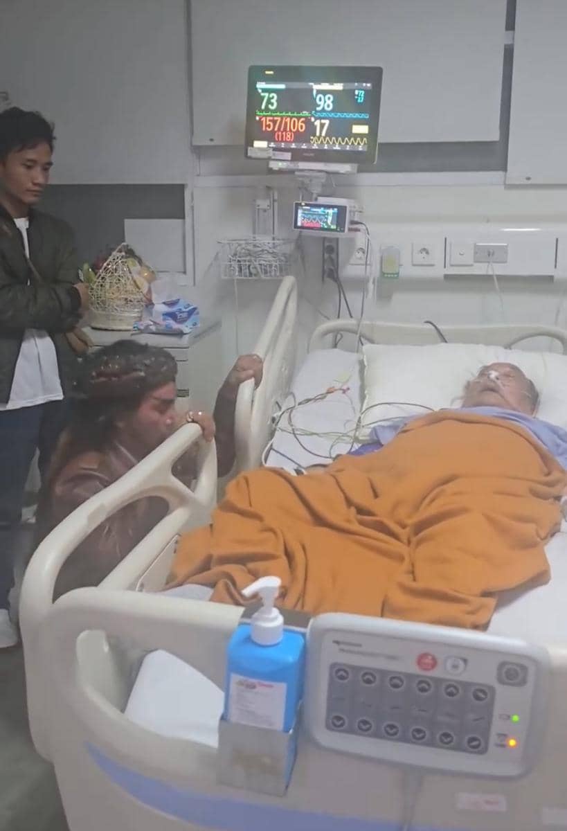 Seorang pria menjenguk pasien lanjut usia yang terbaring di ranjang rumah sakit dengan selimut oranye dan alat medis terpasang.