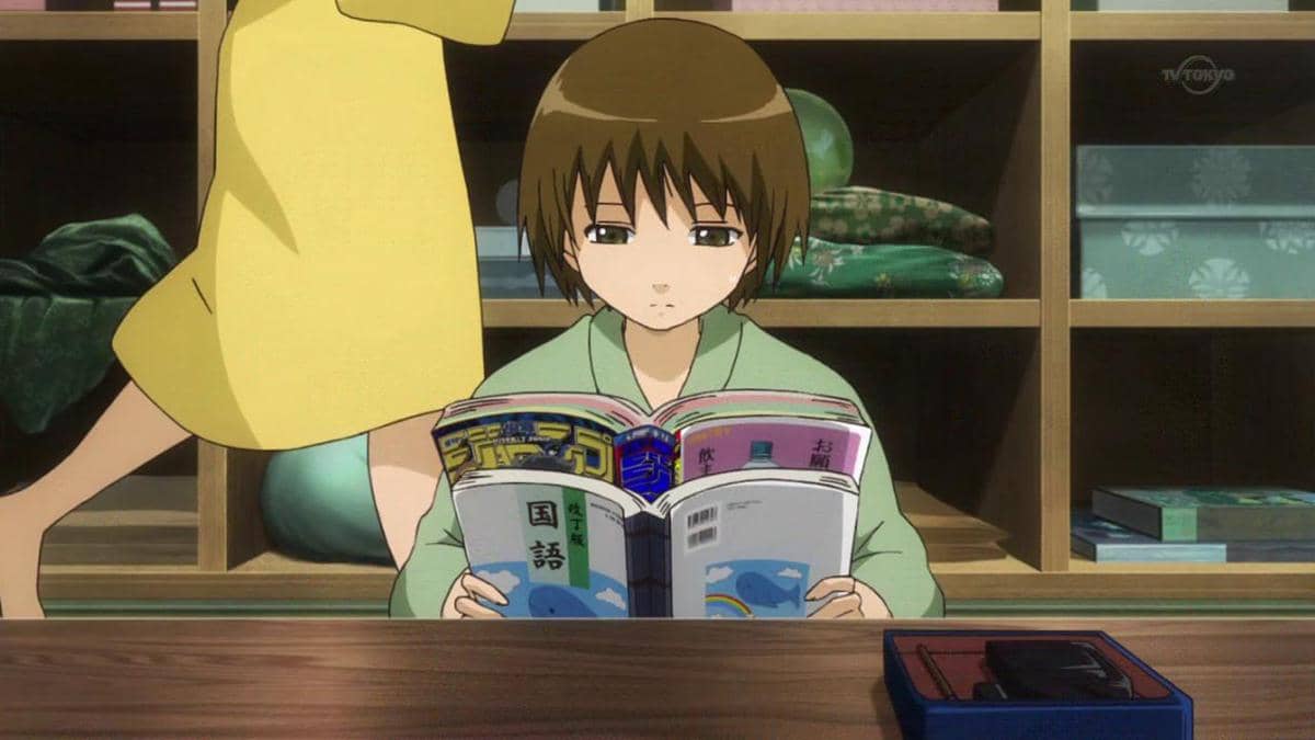 Seita duduk membaca buku di atas meja kayu dengan latar rak berisi bantal dan kotak penyimpanan di ruangan tradisional Jepang.