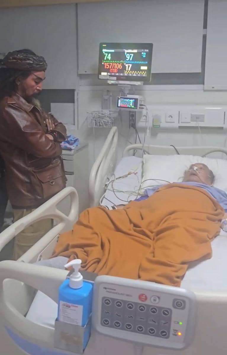 Seorang pria berdiri di samping tempat tidur rumah sakit sambil melihat pasien lanjut usia yang terbaring dengan selimut oranye.