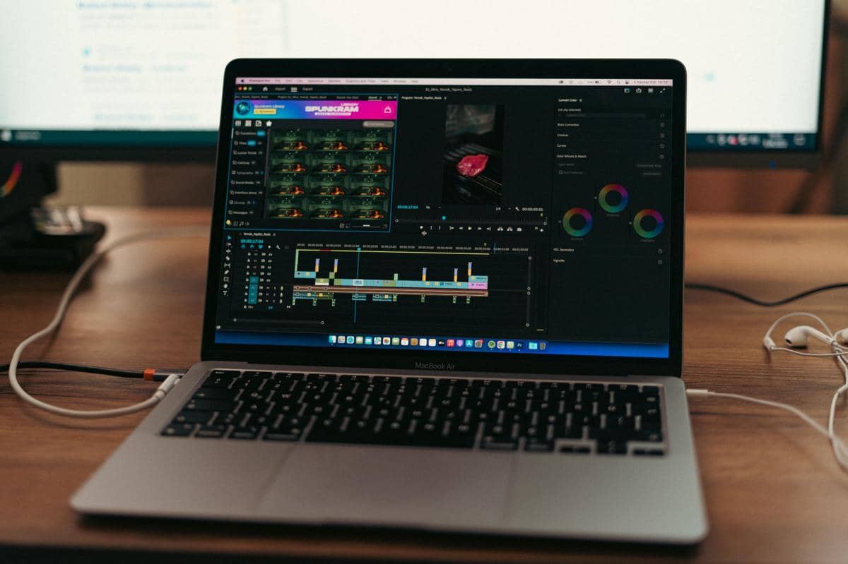 Laptop di atas meja menampilkan perangkat lunak pengeditan video dengan tampilan timeline dan alat koreksi warna di layar.
