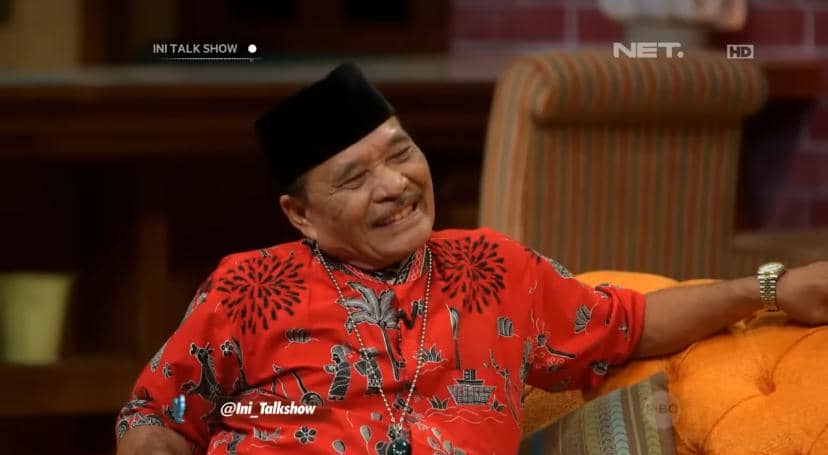 Seorang pria mengenakan baju merah bermotif dan peci hitam sedang tersenyum santai di sofa oranye dalam acara talk show televisi.