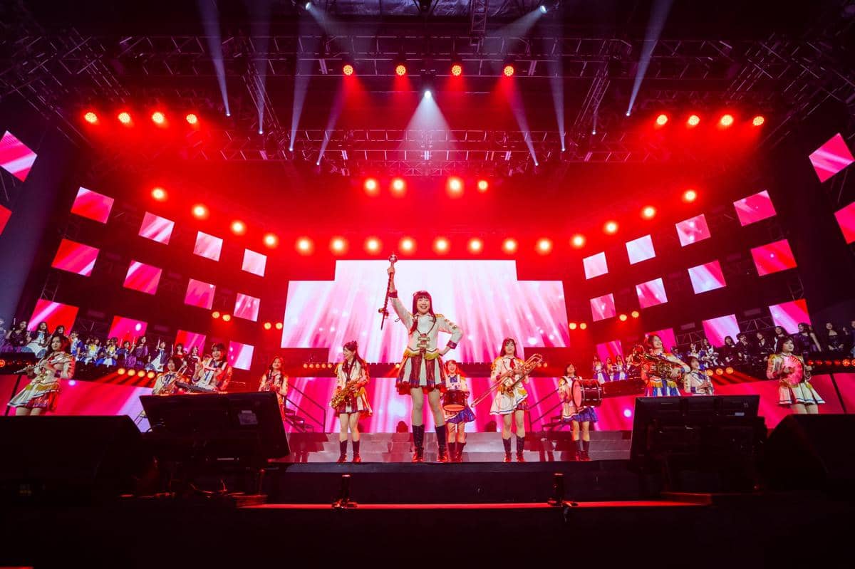 Penampilan lagu Fajar Sang Idola oleh grup idola JKT48 di atas panggung dengan pencahayaan merah dan layar LED besar di latar belakang.