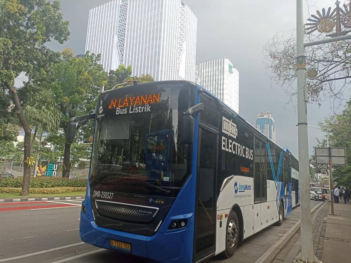 Bus listrik Transjakarta berwarna biru putih berhenti di tepi jalan dengan latar gedung tinggi di kawasan Jakarta.