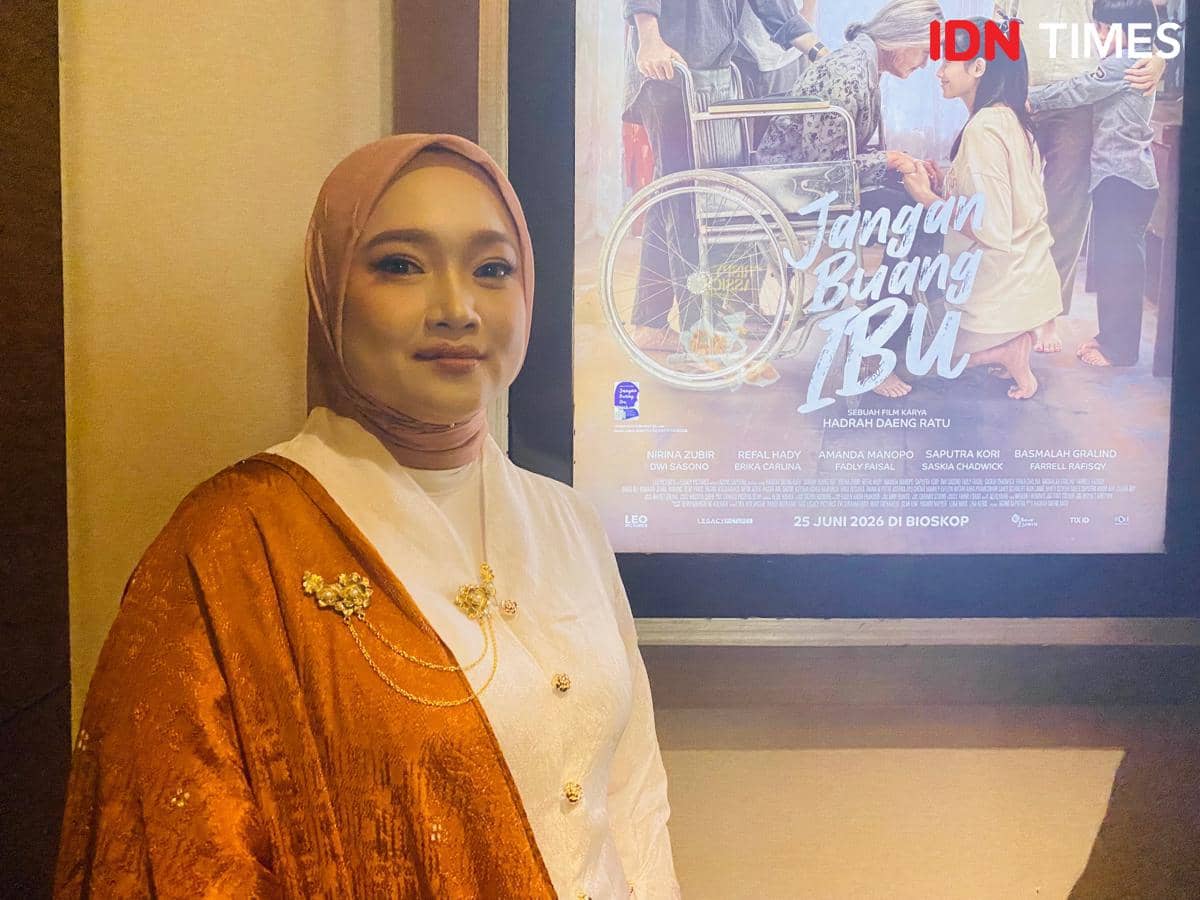 Seorang perempuan berhijab berpose di depan poster film berjudul Jangan Buang Ibu yang menampilkan adegan keluarga dan kursi roda.