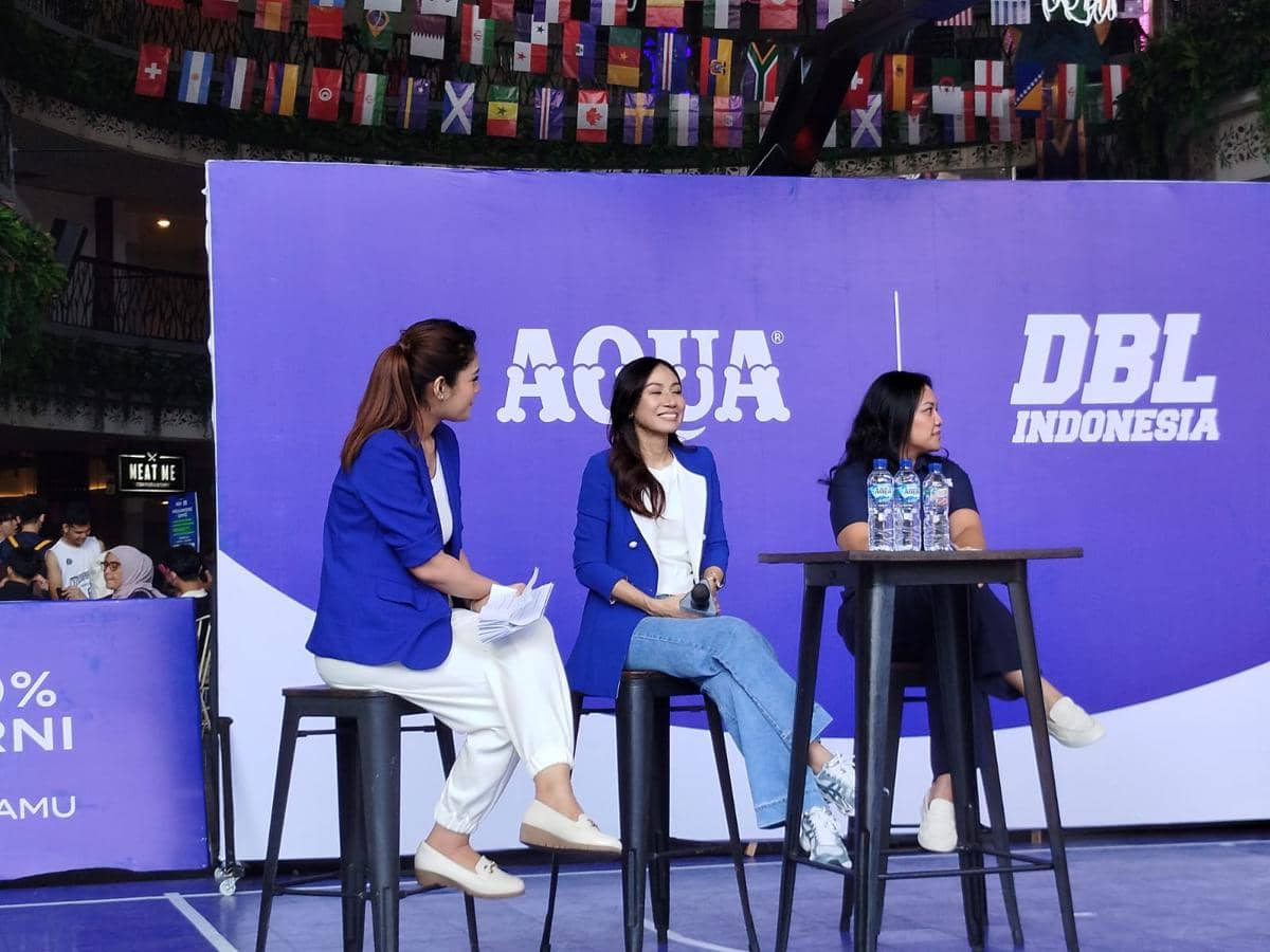 Tiga perempuan duduk di atas panggung dalam acara kolaborasi Aqua dan DBL Indonesia dengan latar berwarna ungu dan logo kedua merek.