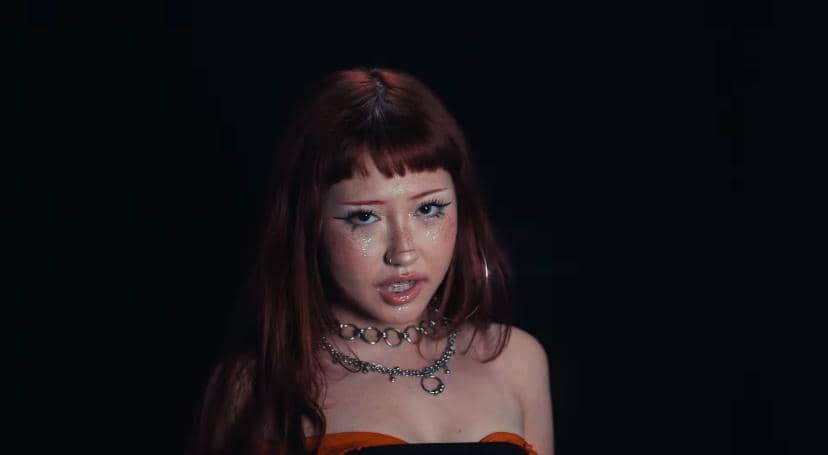 Seorang perempuan dengan riasan berkilau dan kalung rantai tampil di latar gelap dalam cuplikan video klip lagu MMG (My Mine Gueh).
