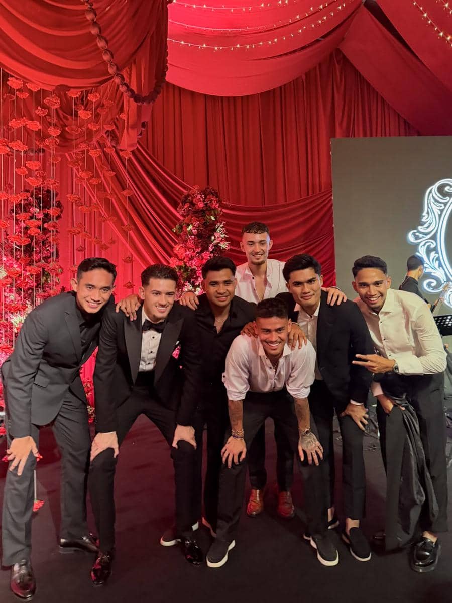 Sekelompok pria berpakaian formal berpose bersama di acara resepsi dengan dekorasi merah dan lampu gantung yang elegan.
