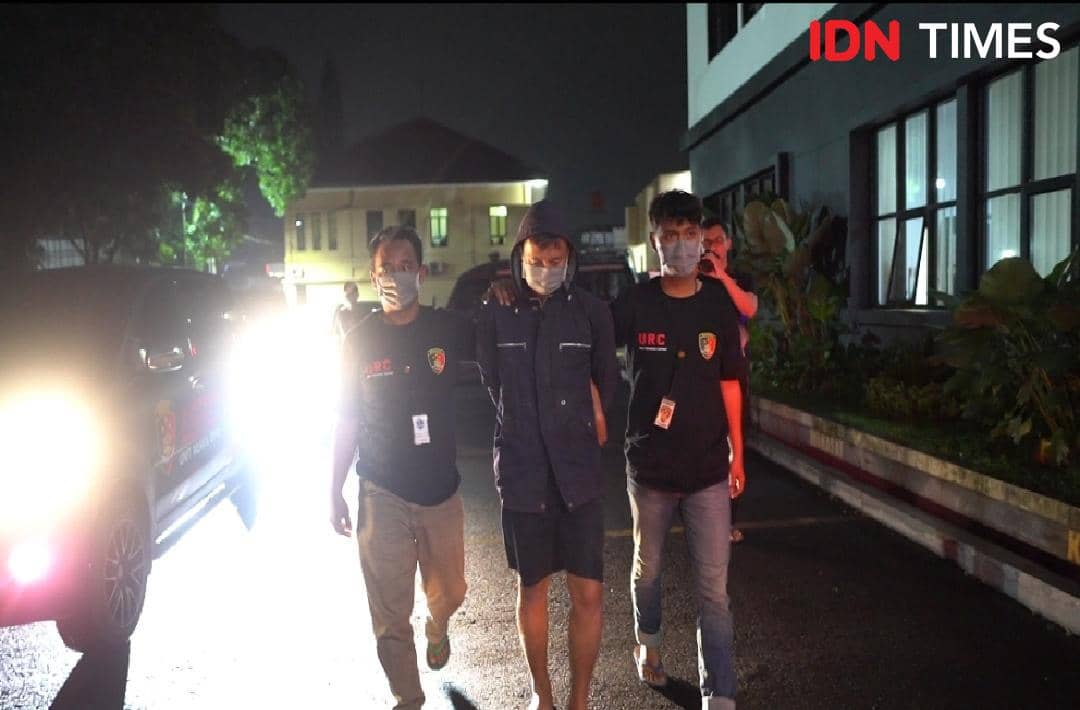 Petugas kepolisian membawa seorang tersangka berpakaian gelap dengan tangan diborgol di area parkir pada malam hari.