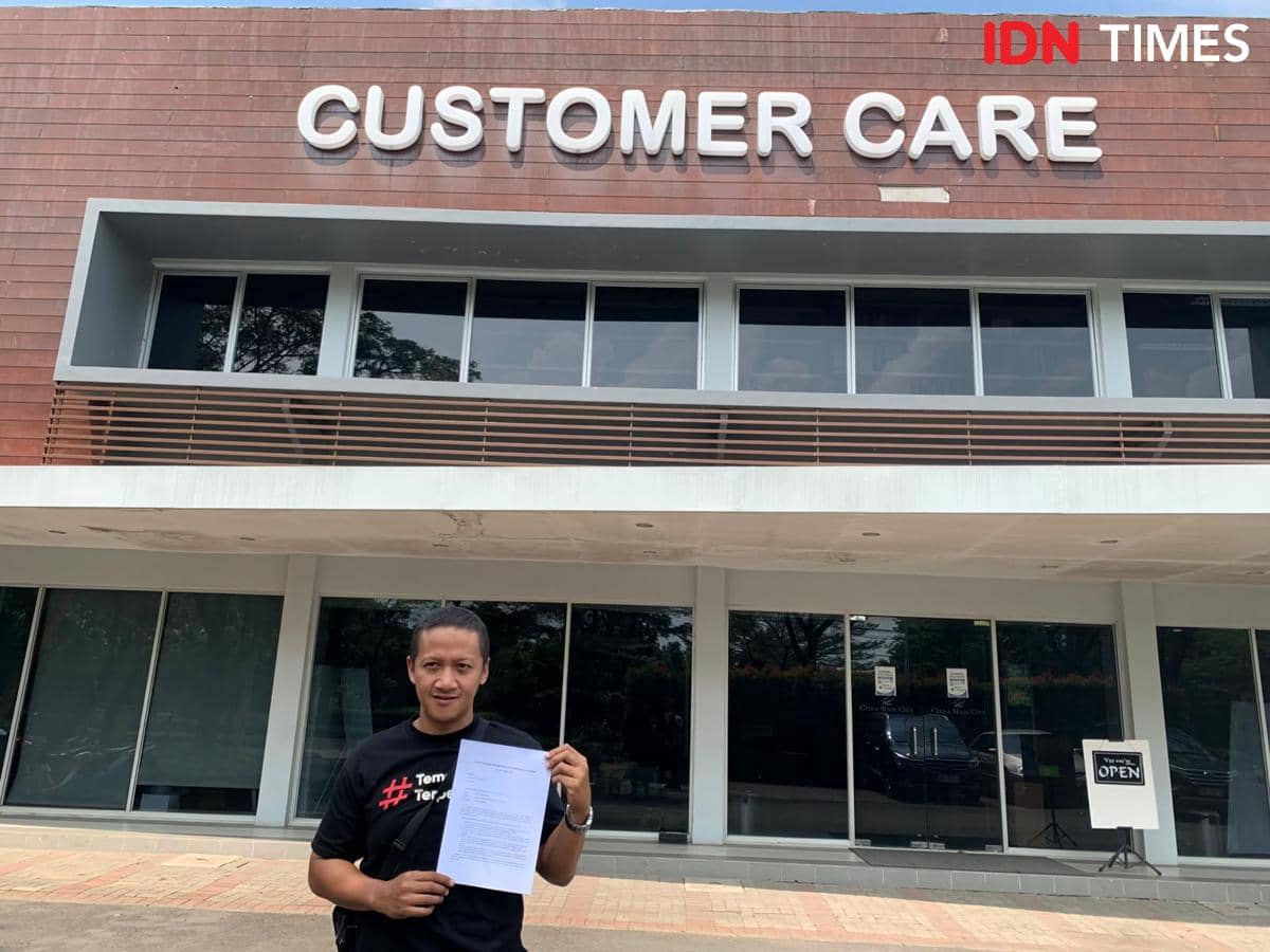 Seorang pria berdiri di depan gedung layanan pelanggan dengan tulisan besar 'Customer Care', memegang selembar dokumen di tangannya.