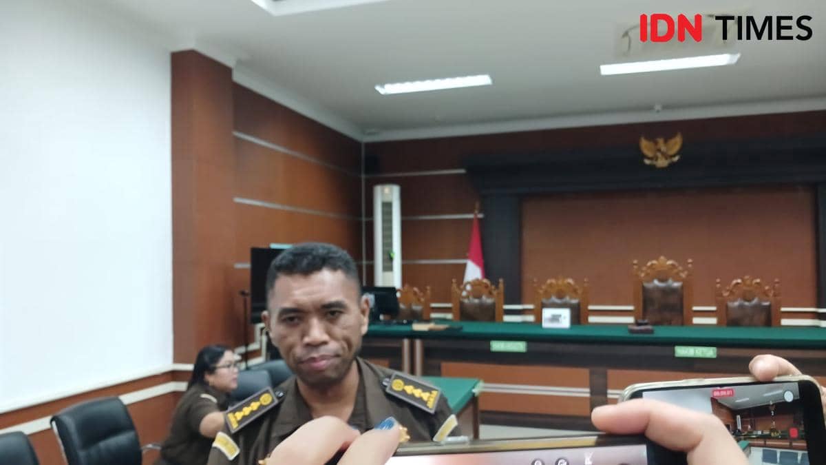 Seorang jaksa berdiri di ruang sidang dengan latar belakang meja hijau, kursi hakim, dan lambang Garuda di dinding belakang.