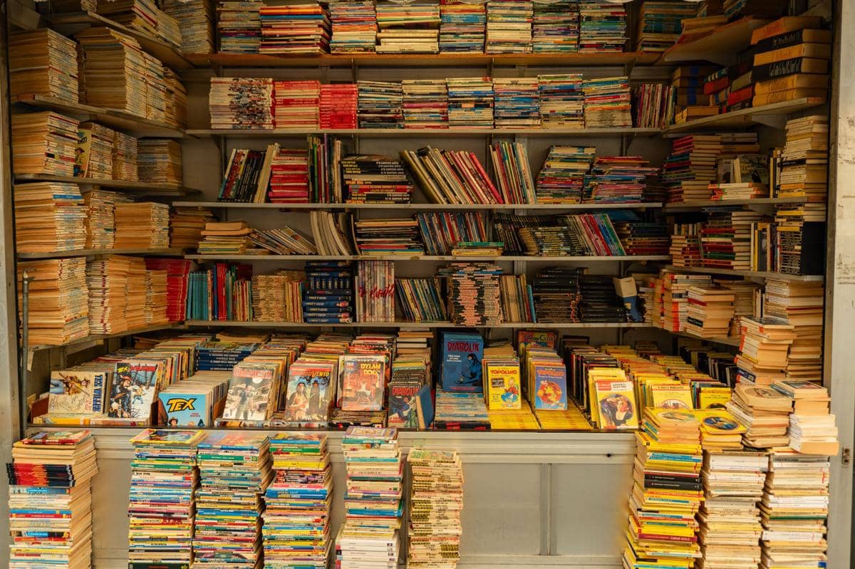 Rak toko buku penuh dengan tumpukan buku dan majalah berwarna-warni yang tersusun rapat dari lantai hingga ke bagian atas rak.