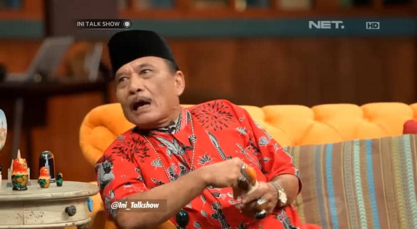 Seorang pria mengenakan baju batik merah dan peci hitam sedang duduk di sofa oranye di acara talk show televisi.