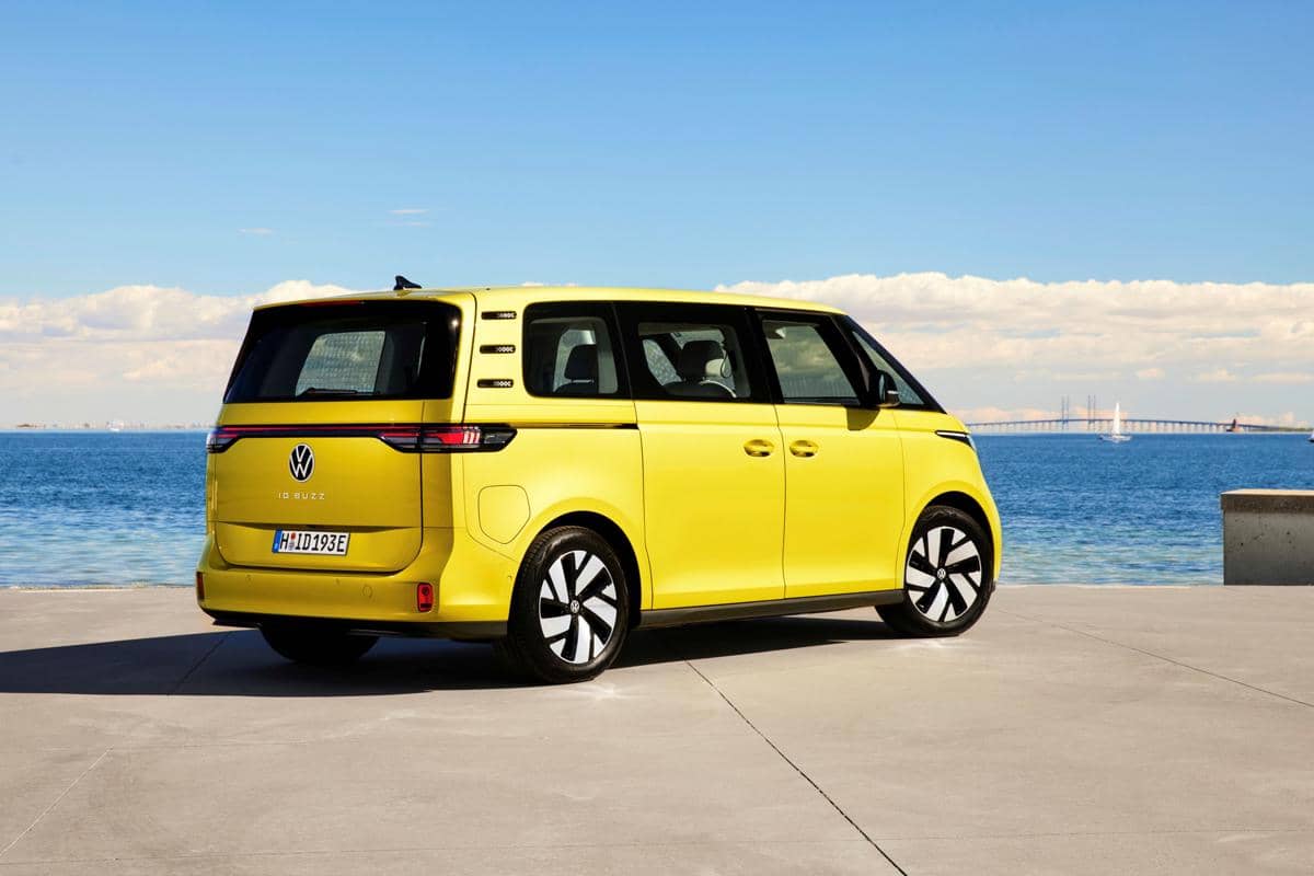 Mobil listrik VW ID. Buzz warna kuning lime terlihat dari belakang diparkir di tepi laut dengan latar langit cerah dan jembatan di kejauhan.