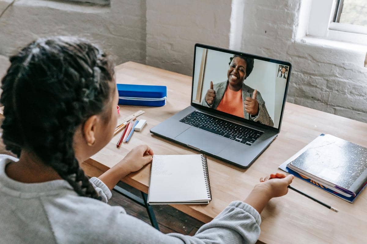 Seorang perempuan mengikuti pertemuan virtual melalui laptop sambil mencatat di buku di atas meja kayu dengan alat tulis di sekitarnya.