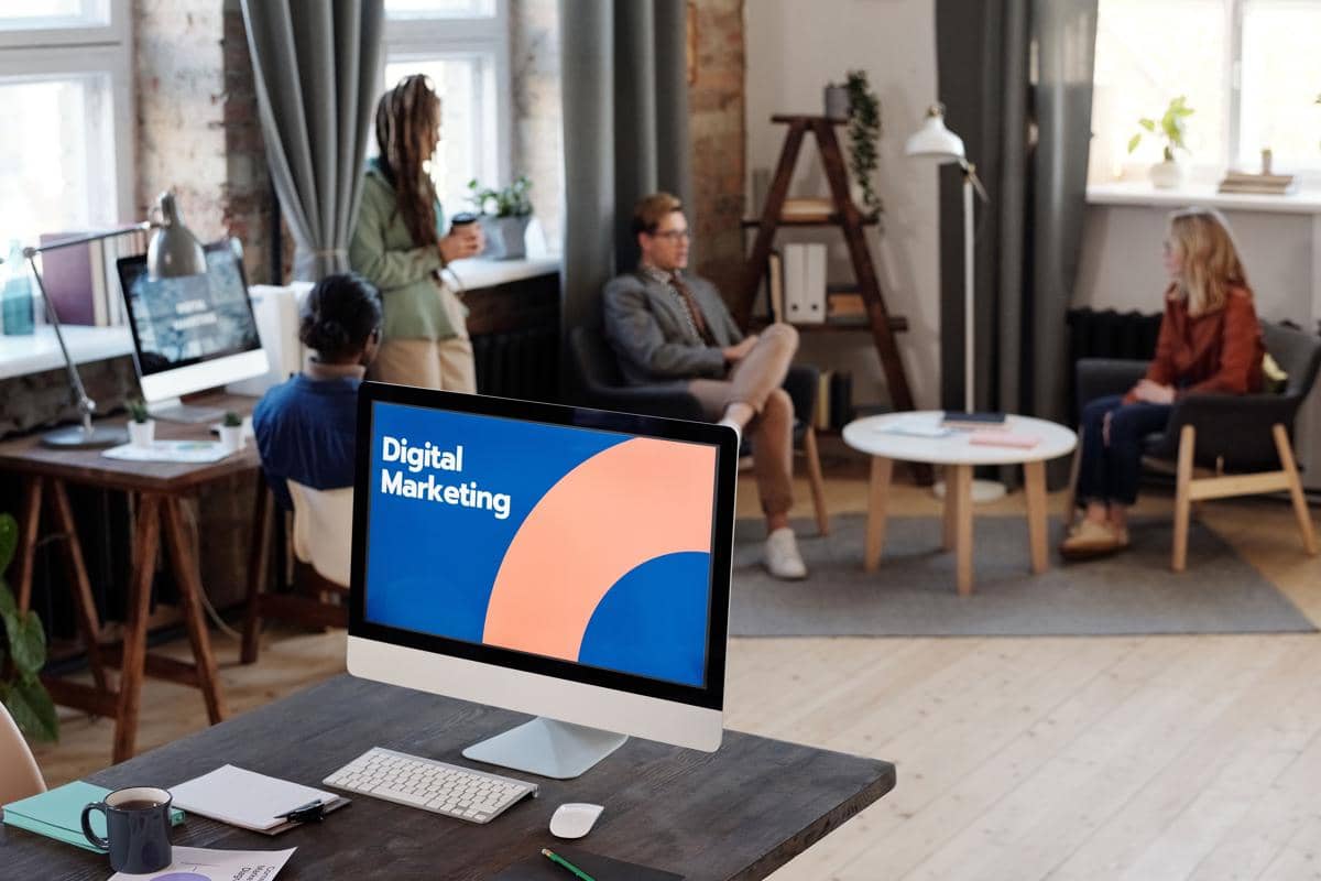 Beberapa orang bekerja dan berdiskusi di kantor modern dengan layar komputer menampilkan tulisan Digital Marketing di meja depan.