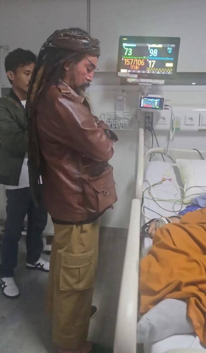 Seorang pria berjaket cokelat berdiri di samping ranjang pasien di rumah sakit, memantau kondisi melalui monitor medis.
